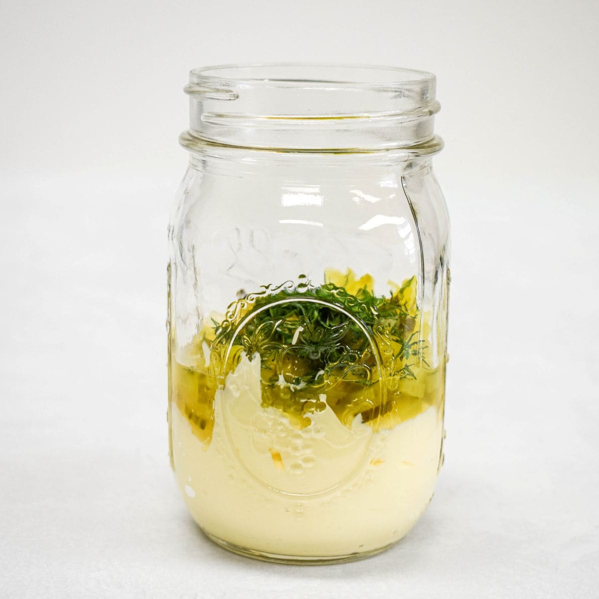 A glass jar filled with mayonnaise, chopped pickles, onion, dill, lemon juice, and seasonings before mixing.