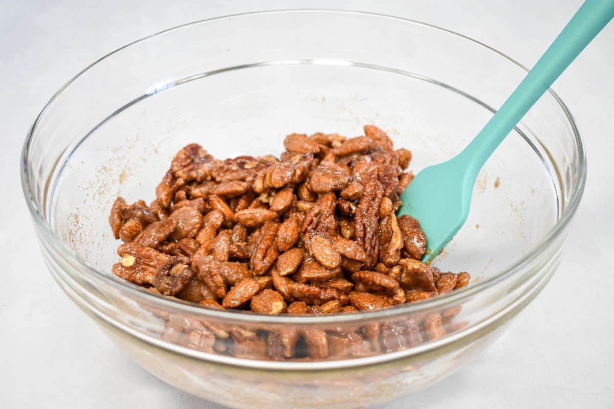Glass bowl filled with nuts fully coated in the sweet spiced mixture, with a teal spatula.
