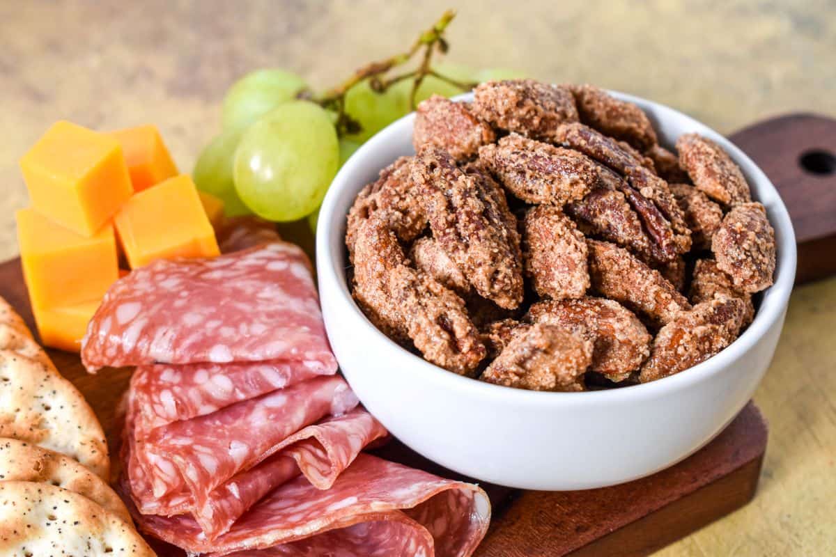 Sweet spiced nuts mix served in a white bowl with cheese, salami, grapes, and crackers on a wooden board.