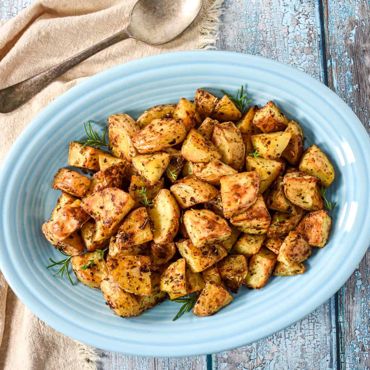 Roasted gold potatoes served on a blue platter with a spoon and cloth on a rustic wood table.