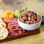 White bowl filled with sweet spiced nuts next to sliced salami, cheddar cheese cubes, crackers, and green grapes on a wooden serving board.