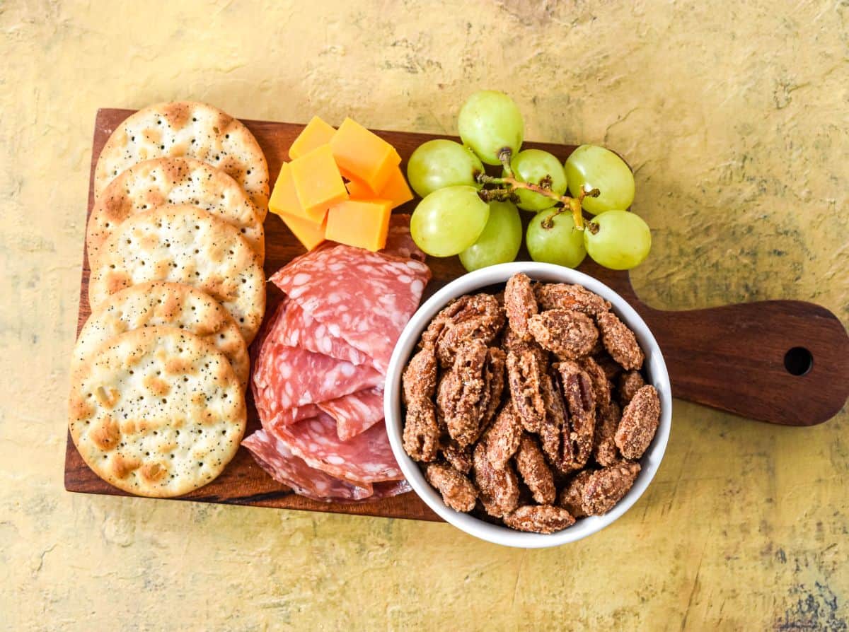 Top-down view of sweet spiced nuts in a white bowl surrounded by crackers, salami, cheese cubes, and green grapes on a wooden board.