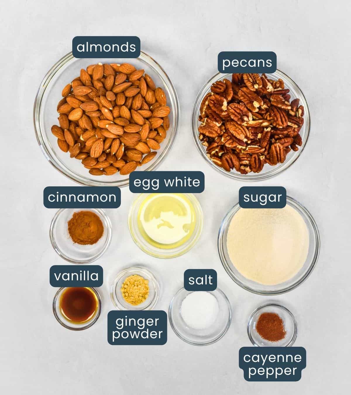 All recipe ingredients laid out in glass bowls and labeled: almonds, pecans, egg white, sugar, cinnamon, salt, ginger powder, cayenne pepper, and vanilla.