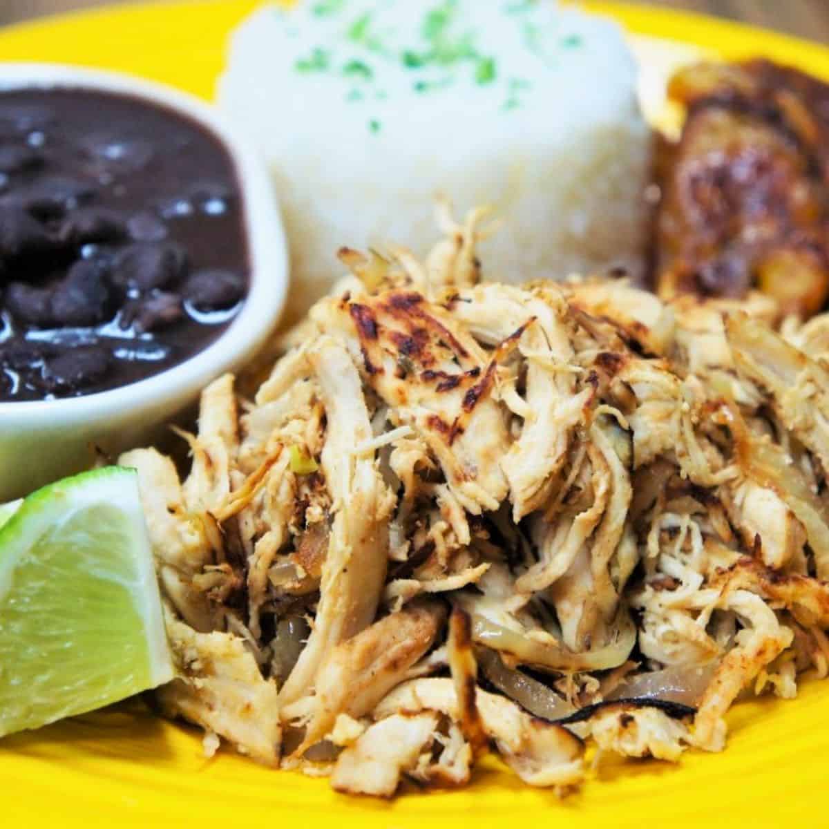 Shredded chicken served with white rice, black beans, plantains and lime wedges on a yellow plate.