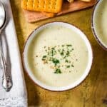 Bowl of creamy celery soup garnished with chopped parsley and black pepper, served with crackers on a wooden board.