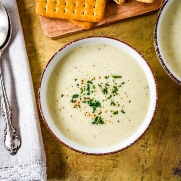 Bowl of creamy celery soup garnished with chopped parsley and black pepper, served with crackers on a wooden board.