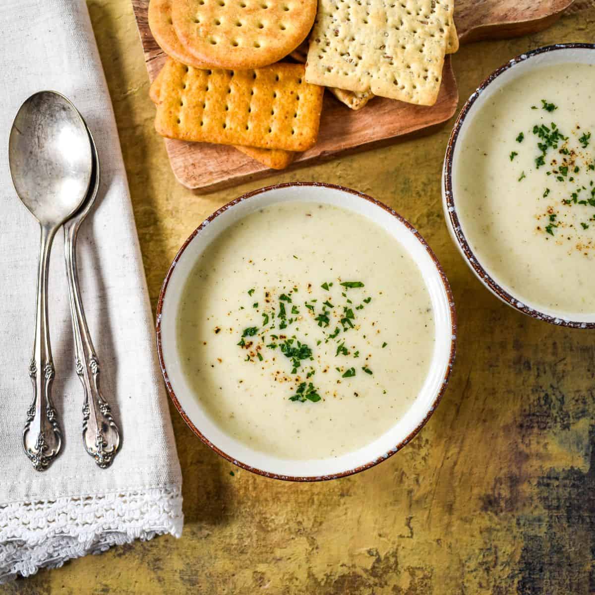 Two bowls of cream of celery soup topped with parsley and pepper, next to a stack of crackers and silver spoons on a rustic table.