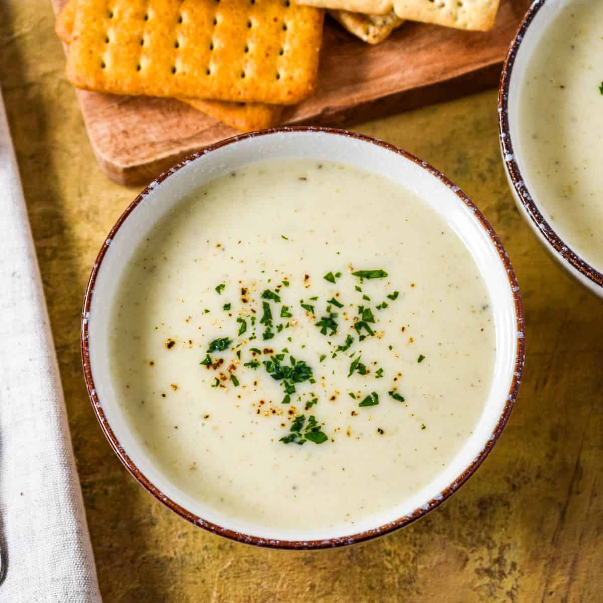 Bowl of creamy celery soup garnished with chopped parsley and black pepper, served with crackers on a wooden board.