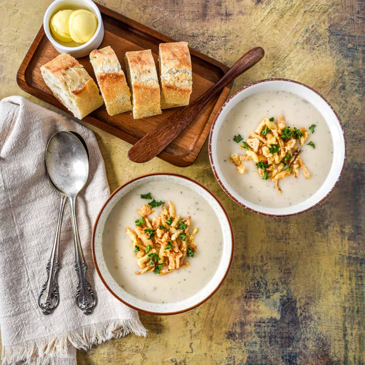 Two white bowls of creamy onion soup topped with crispy fried onions and parsley, served with sliced bread and butter.