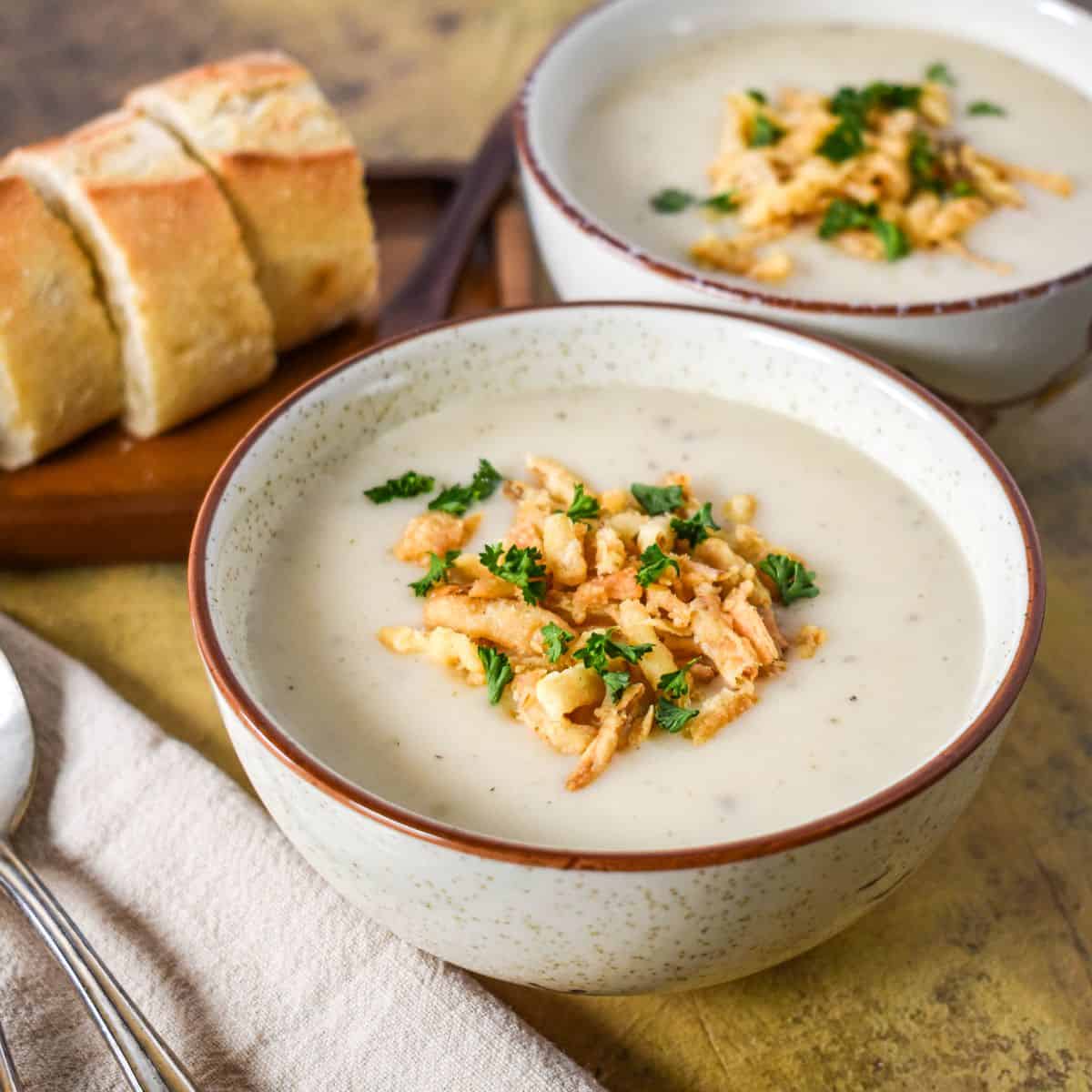 Close-up of creamy onion soup in a white bowl, topped with crispy fried onions and fresh parsley, with bread in the background.