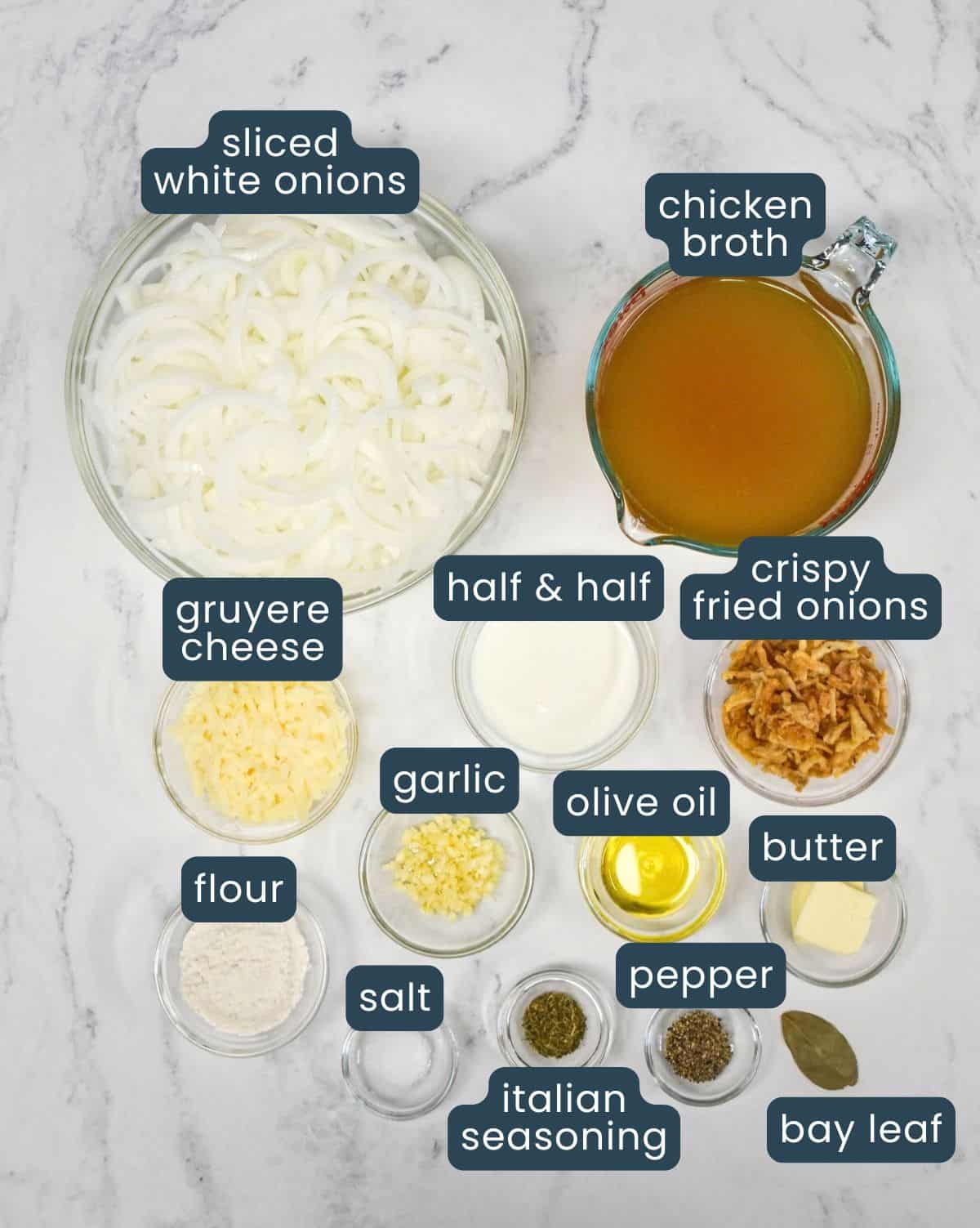 Ingredients for cream of onion soup, prepped and arranged in glass bowls on a white surface, including white onions, chicken broth, Gruyere cheese, half-and-half, crispy fried onions, garlic, butter, olive oil, flour, and seasonings.