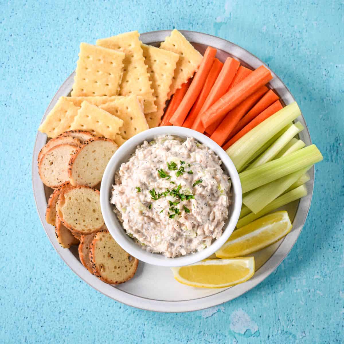 Salmon salad served in a bowl surrounded by crackers, bagel chips, carrot sticks, celery, and lemon wedges.