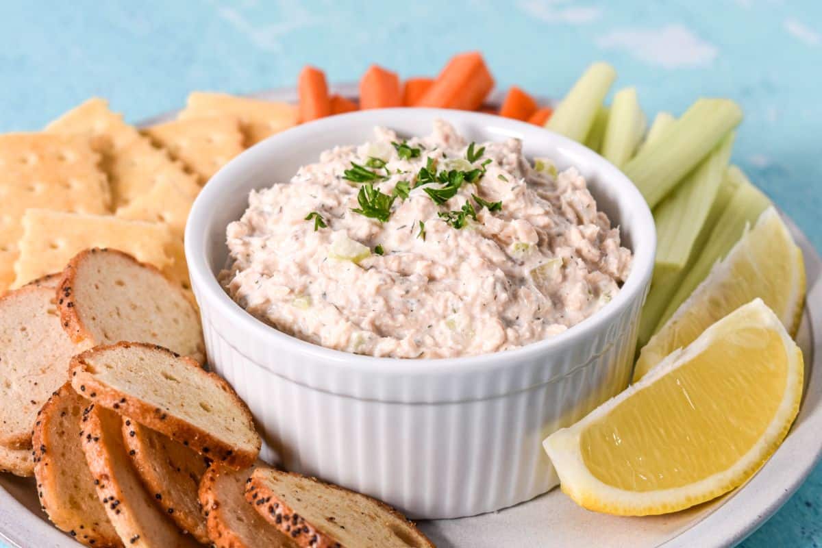 Close-up of creamy salmon salad with celery and dill, topped with chopped parsley and served with bagel chips and lemon.
