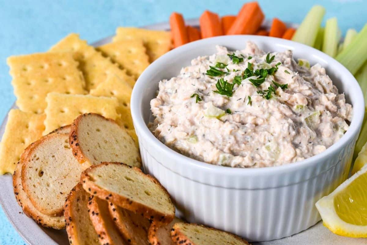 Creamy salmon salad garnished with parsley and served with bagel chips, crackers, fresh vegetables, and lemon.