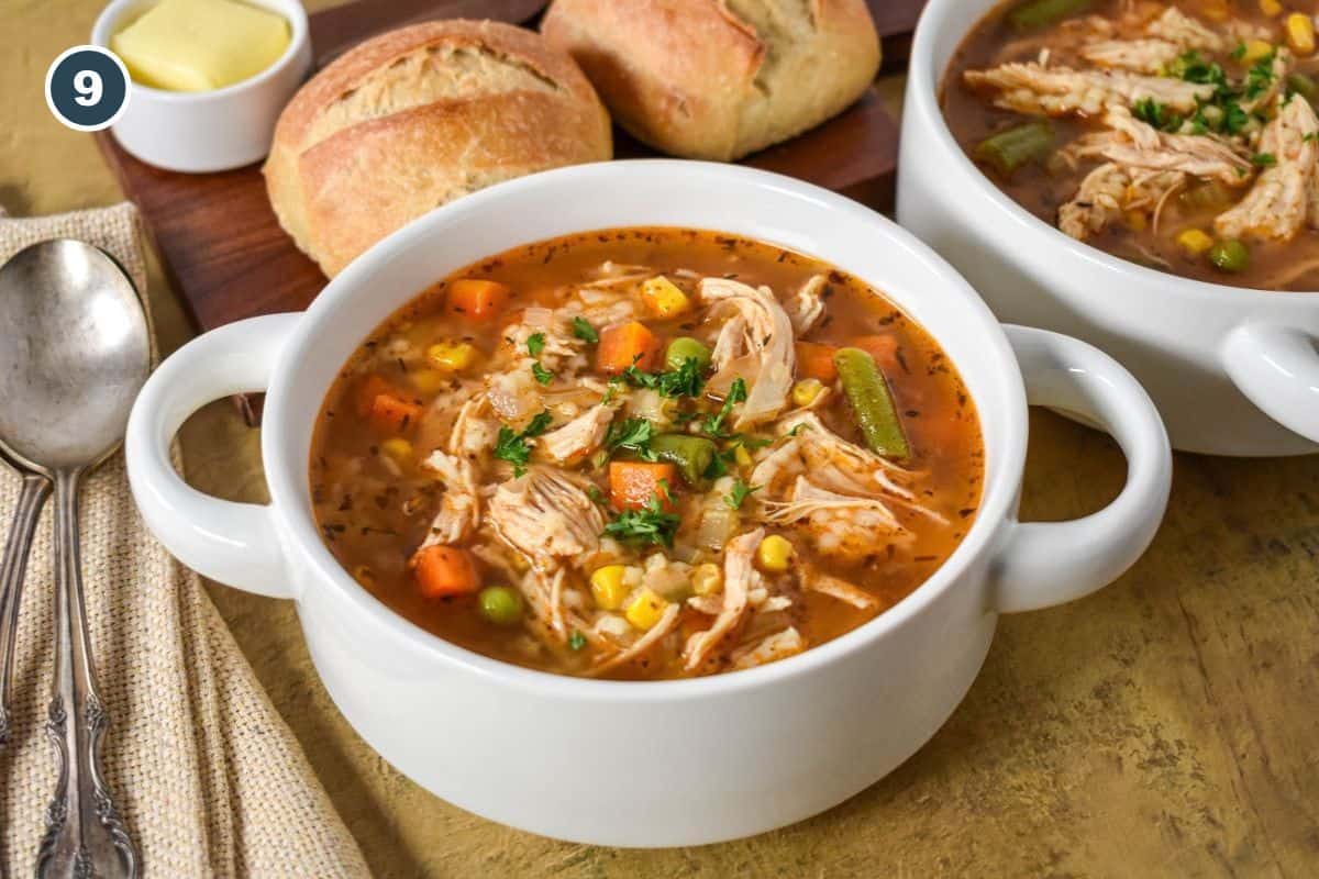 Finished chicken vegetable soup with shredded chicken, mixed vegetables, and pastina in a flavorful broth, served with bread.
